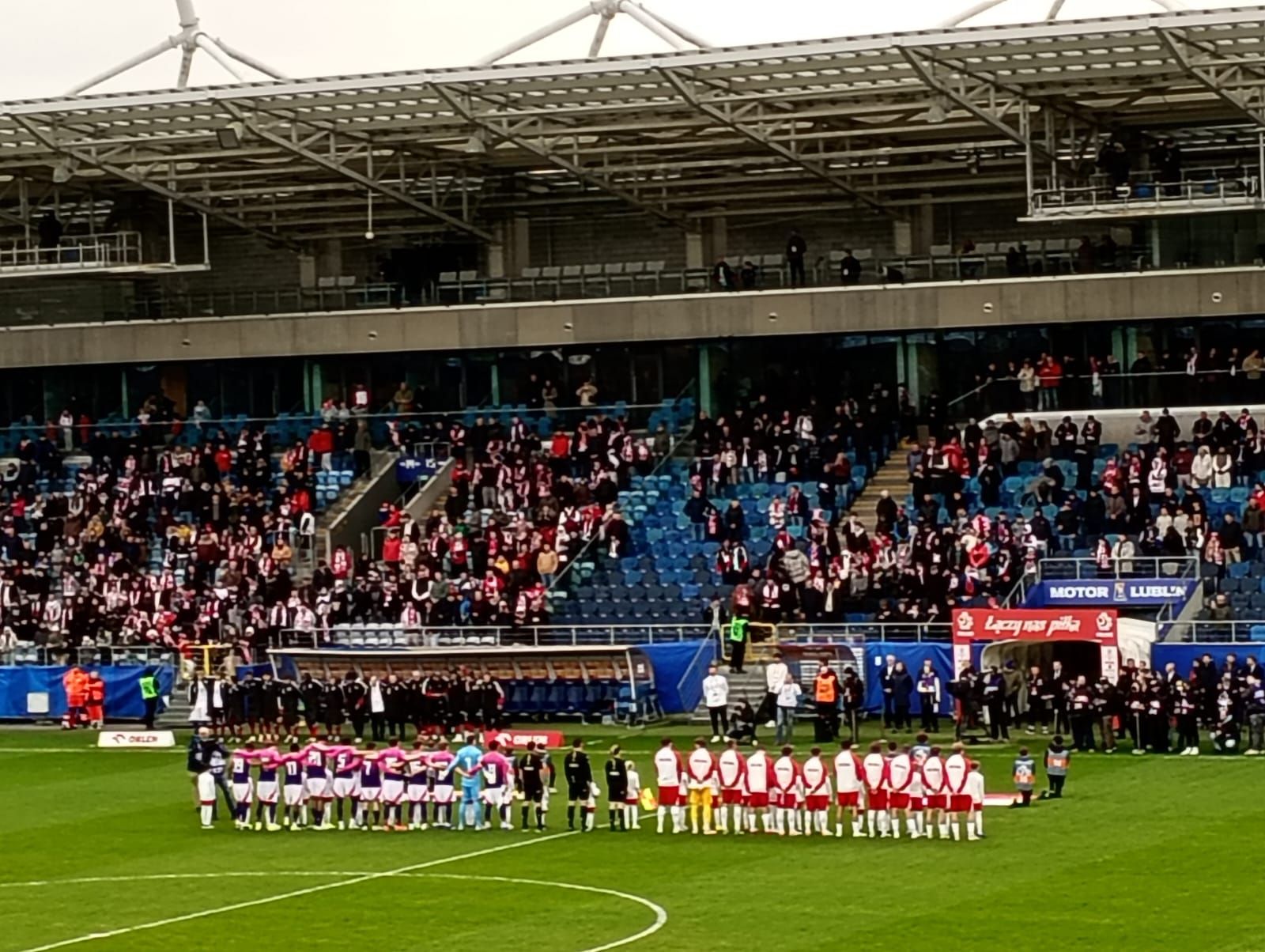 Zawodnicy obu drużyn ustawieni na boisku przed rozpoczęciem meczu, w tle trybuny z kibicami.